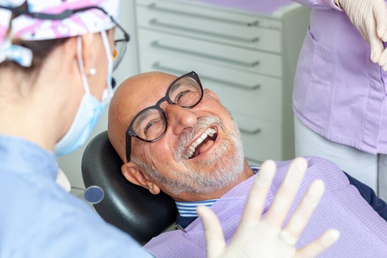 An older man with glasses smiles and laughs in a dentist chair, wearing a bib. A dental professional in a mask and gloves speaks to him, with another person in scrubs standing nearby.