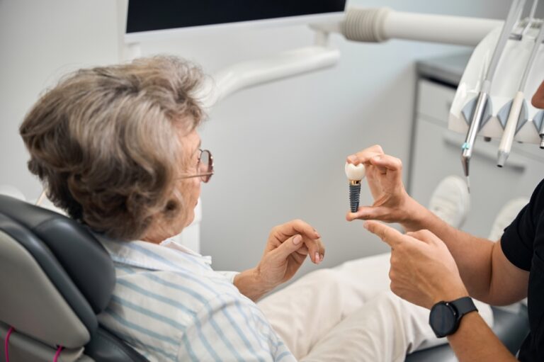 An elderly woman sits in a dental chair as a dentist shows her a model of a dental implant and explains the procedure. Dental equipment and a monitor appear in the background.